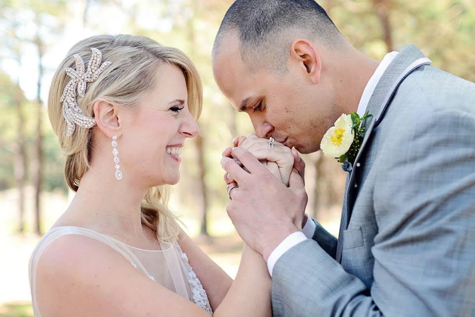 A groom, dapper in his gray suit and yellow boutonnierre, kisses the hands of his smiling bride.