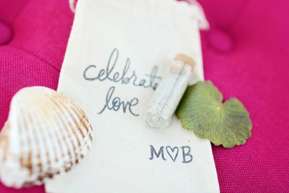A bag embroidered with "celebrate love" lies on a fuchsia pink couch with a vial of sand and a shell to commemorate the bride and groom's beach wedding.