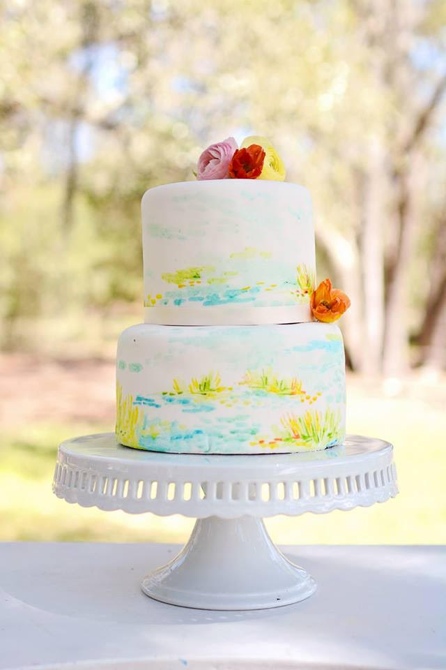 A two-tiered cake hand-painted with blue and green watercolors rests atop a milk glass cake stand.