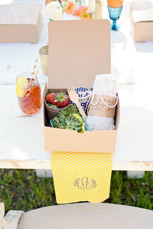A wrapped sandwich and strawberries fill a craft paper food box that sits on a monogrammed napkin next to a mason jar full of sweet tea.