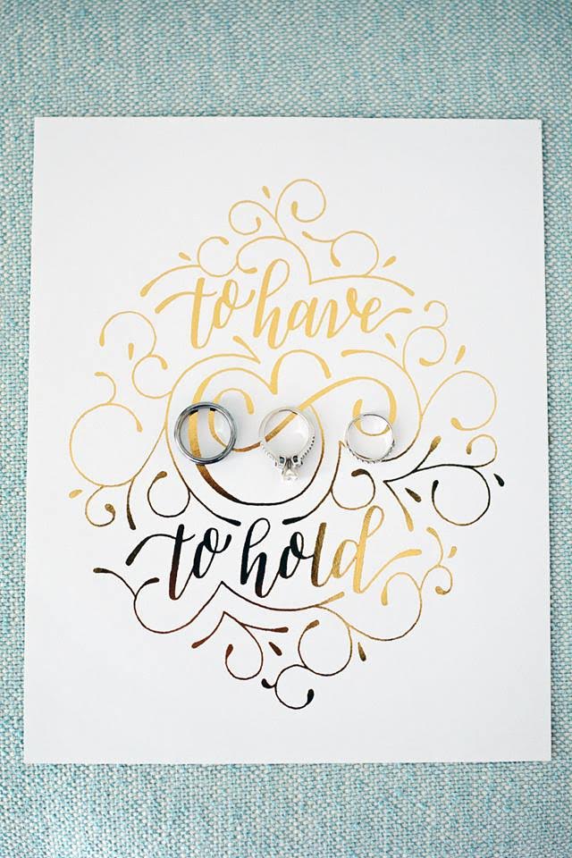 The bride's wedding and engagement rings sit next to her groom's wedding band on top of a gold scripted card reading "to have and to hold."