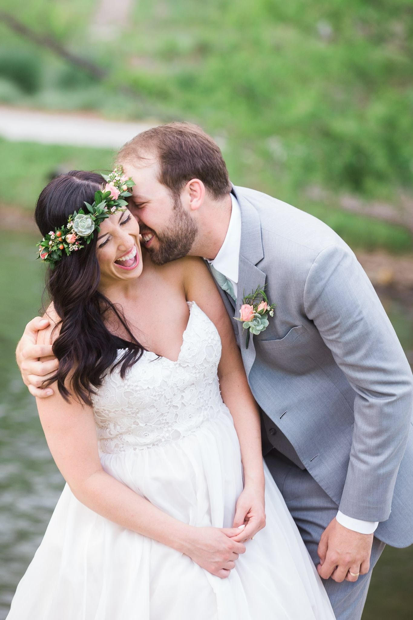 Bride wearing a flower crown over her loosely curled hair style laughs as her groom nuzzles into her ear.