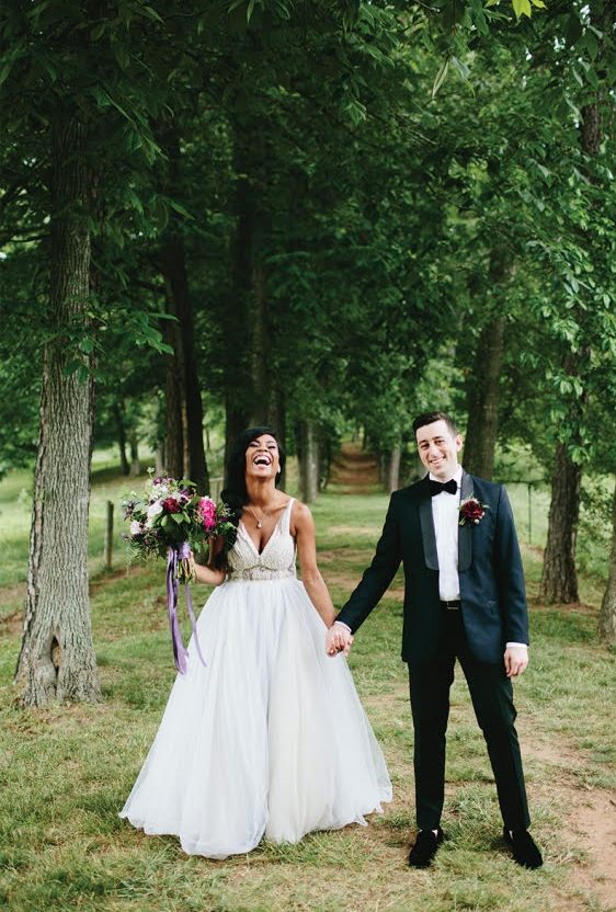 Bride and groom laugh as they hold hands standing outside in a grove of trees.