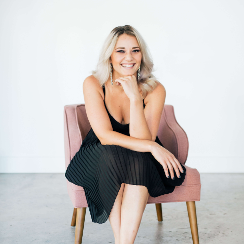 Asheville hair stylist Zhenya Lazarchuk smiles as she sits in a blush pink armchair with her chin resting on her fingers.