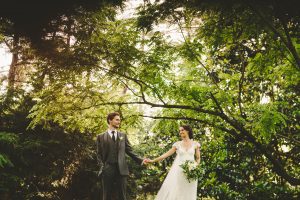 Bride and groom stand apart while holding hands under green leafy tree branches.