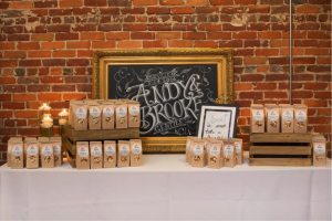 A chalkboard sign reading "Andy and Brooke" sits against a brick wall behind paper bags of popcorn at a wedding reception.