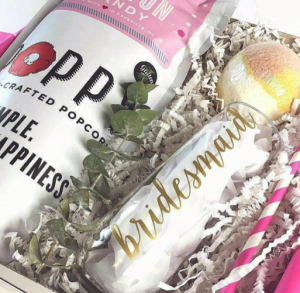 A bag of cotton candy Poppy Handcrafted Popcorn, a tall tumbler that says "bridesmaid", and a pink swirly straw sit atop silver confetti in a thank you gift box.