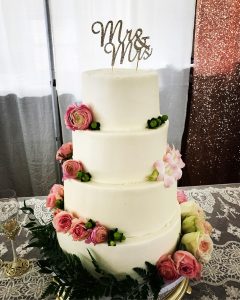 A four-tier ivory wedding cake with pink roses and a filigree Mr and Mrs cake topper sits on a table in front of a window.